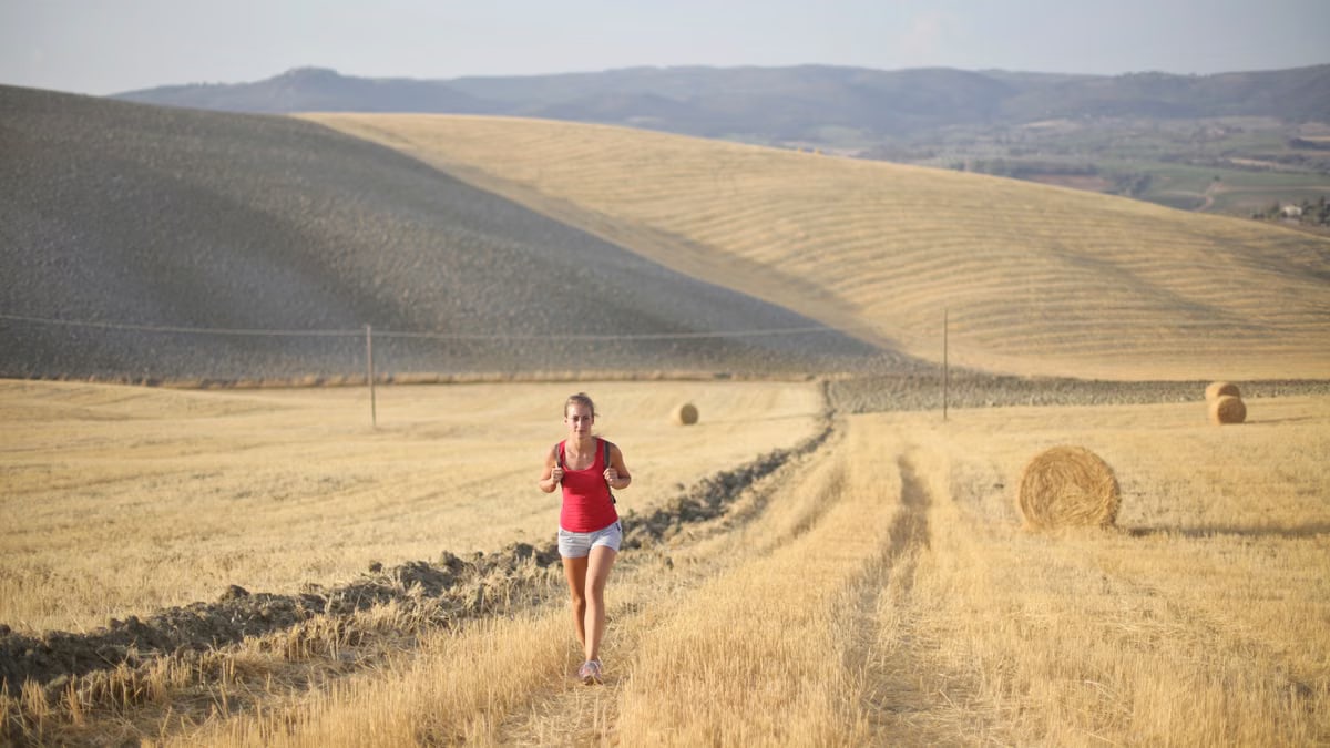 A woman jogs through a scenic rural hayfield on a sunny day, surrounded by rolling hills and hay bales.